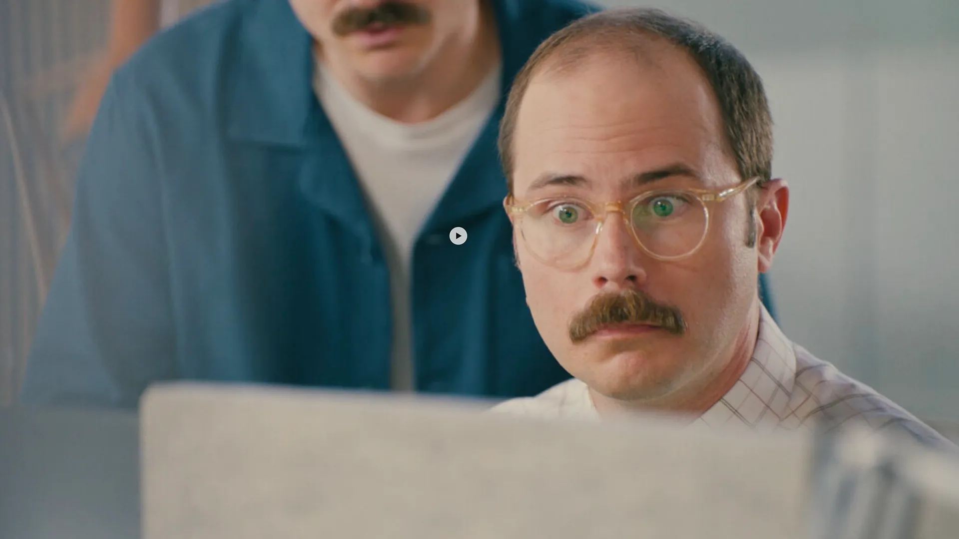 Close-up of a wide-eyed, mustached man in glasses staring at a laptop screen while another man leans in over his shoulder, both reacting to something on the display