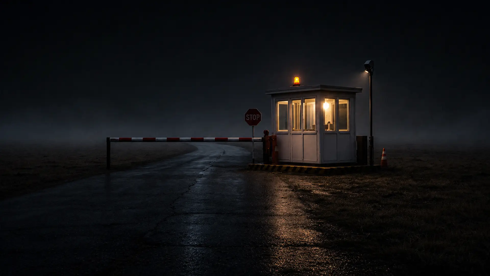 A lit security checkpoint booth beside a lowered red and white barrier arm on a wet road at night, with a stop sign and an amber beacon glowing through fog