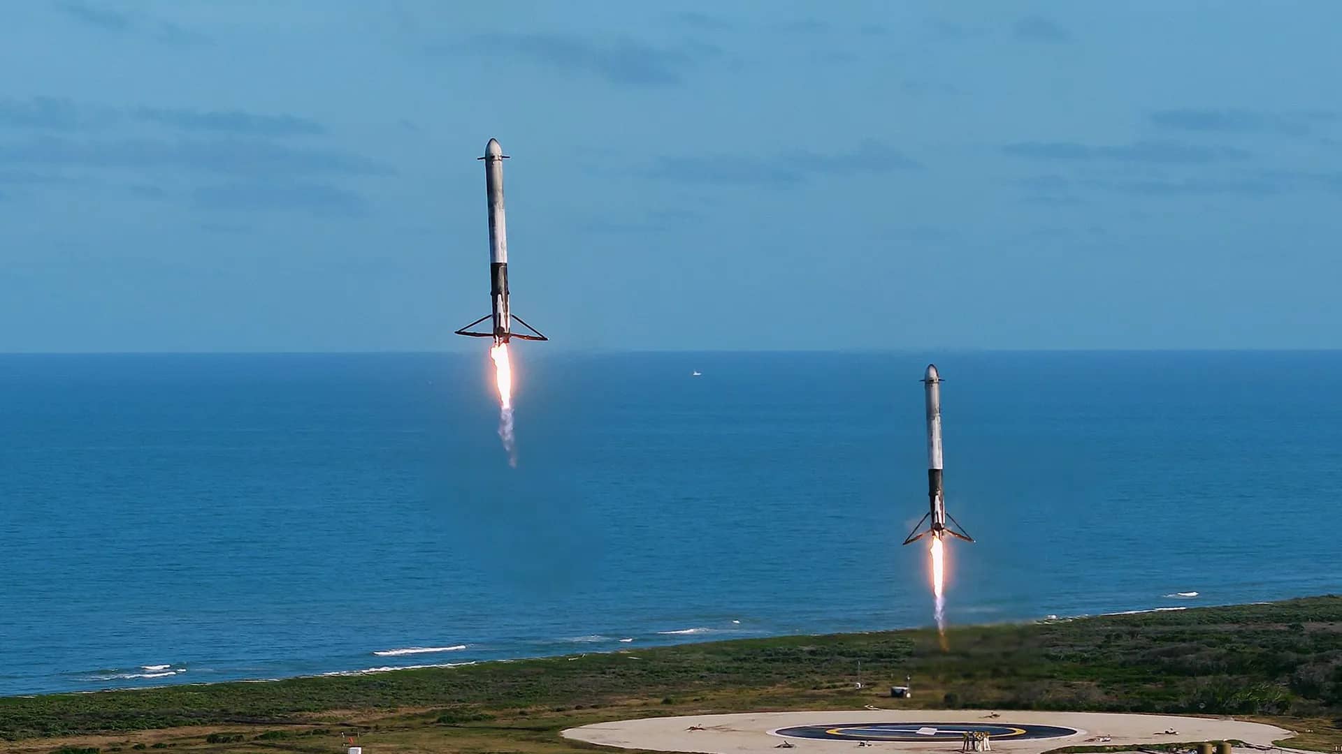 Two reusable rocket boosters descending with engine flames toward a coastal landing pad, framed by vast open ocean and sky that surround the subjects with empty space
