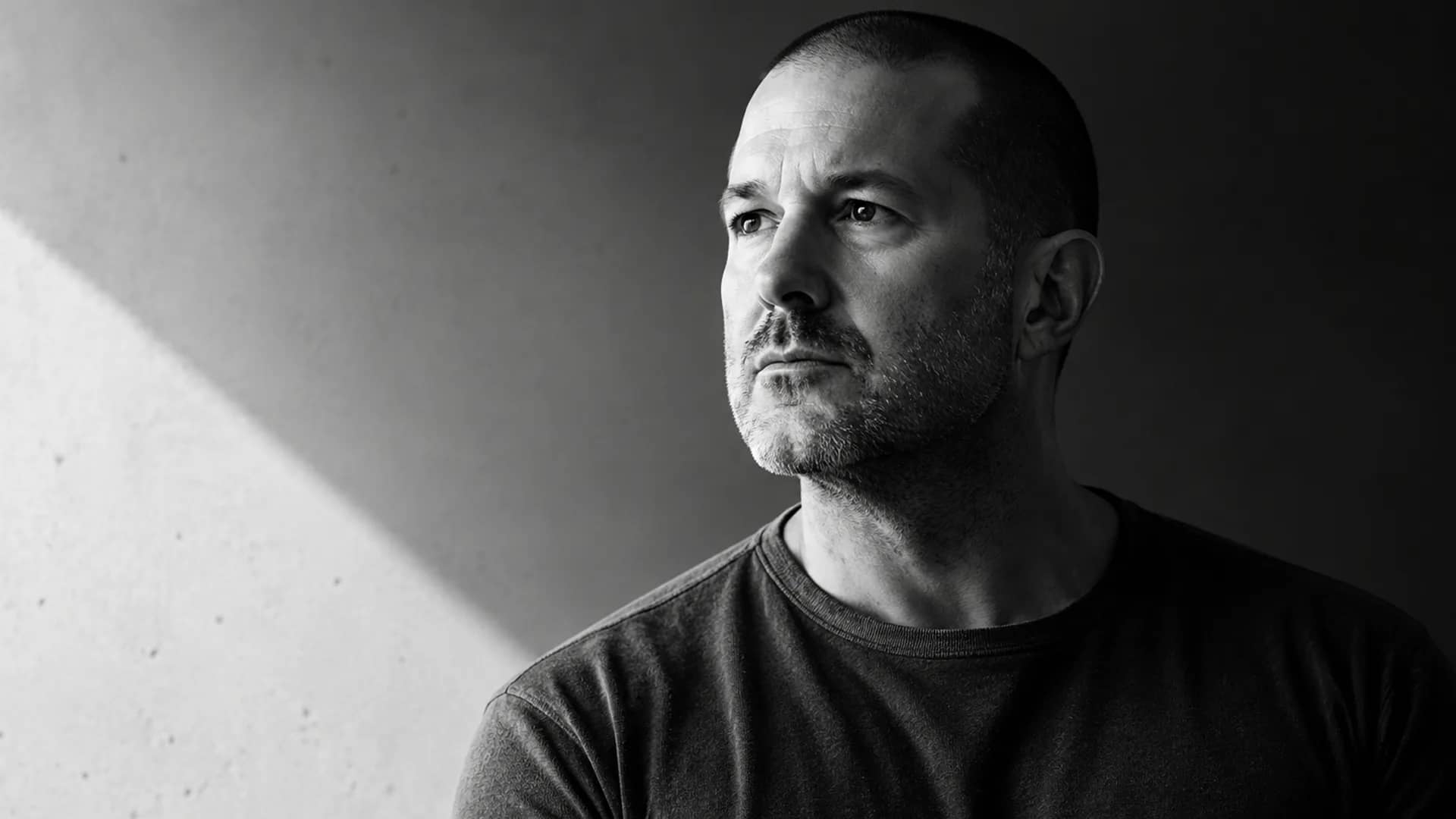 Black-and-white portrait of a bald, stubbled man in a dark crew-neck t-shirt gazing off-frame, a diagonal shaft of light cutting across the shadowed concrete wall behind him